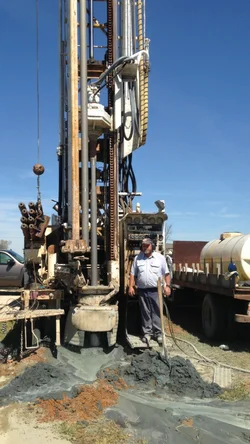 Danny Wright, owner and third-generation driller, standing at the drill rig with gray rock cuttings piled on red clay